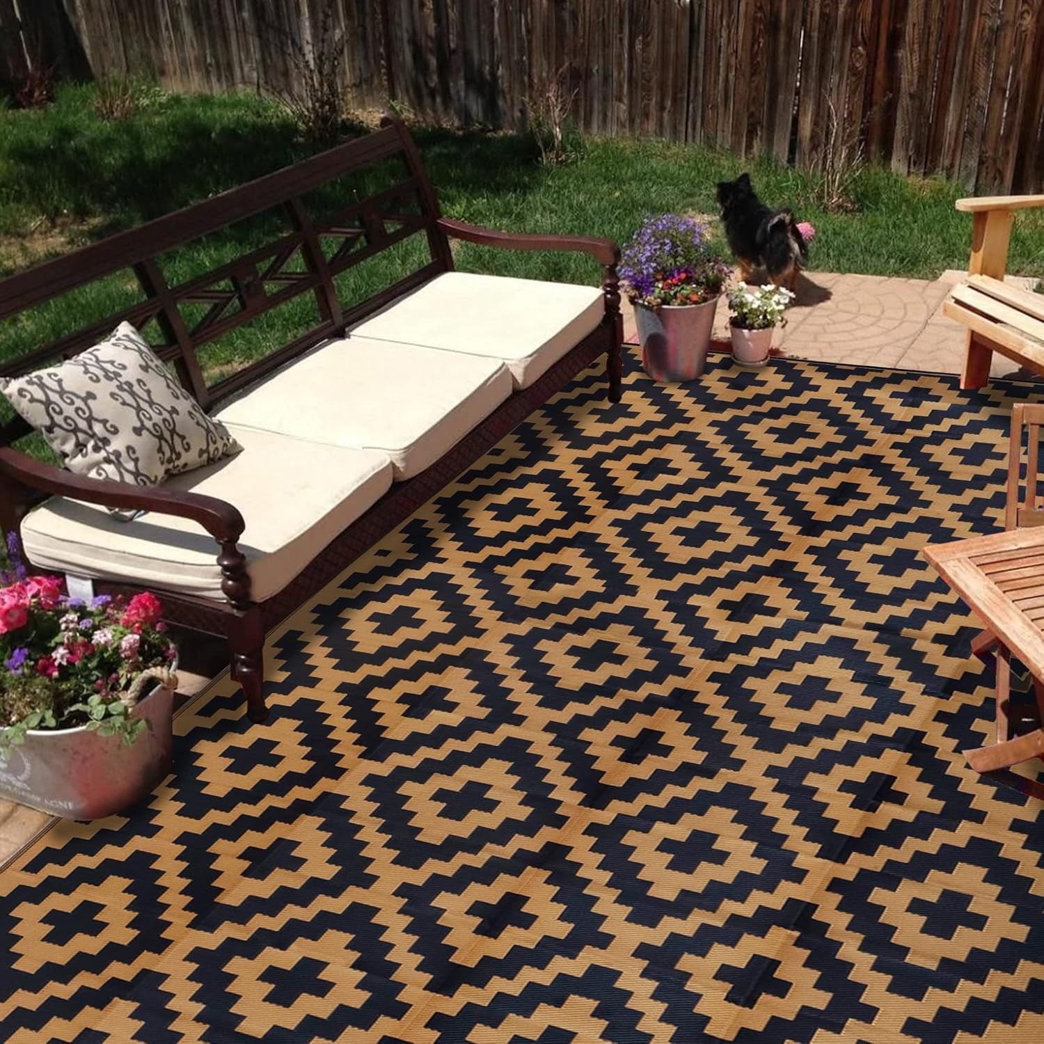 Tan-and-black geometric outdoor rug on a patio under a wooden bench and potted flowers