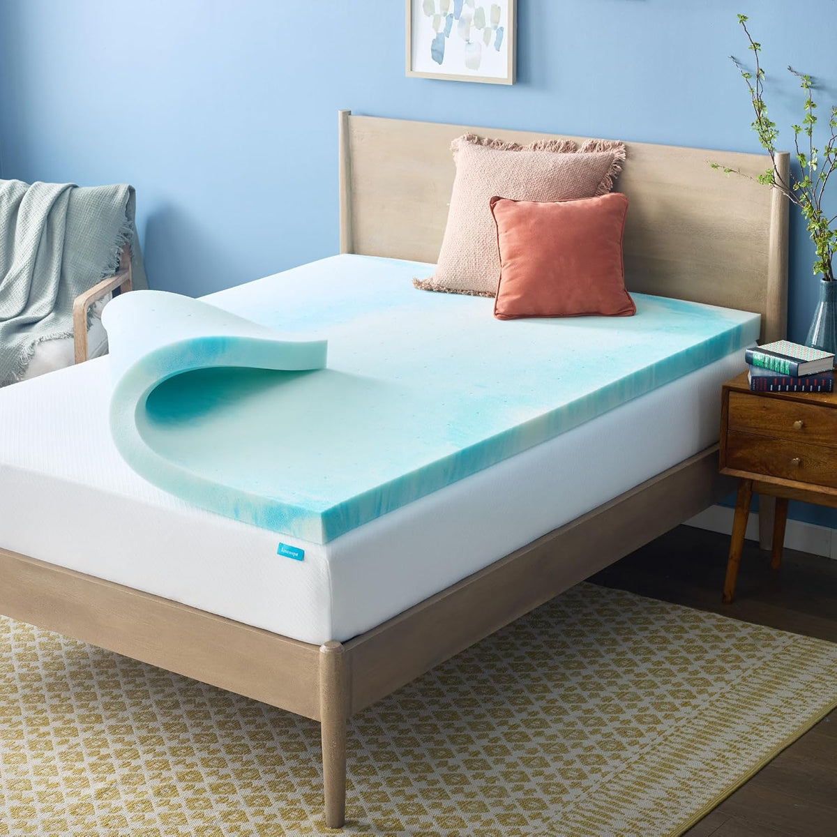 Bedroom scene with a light blue foam topper partially rolled on a mattress and pillows at the headboard