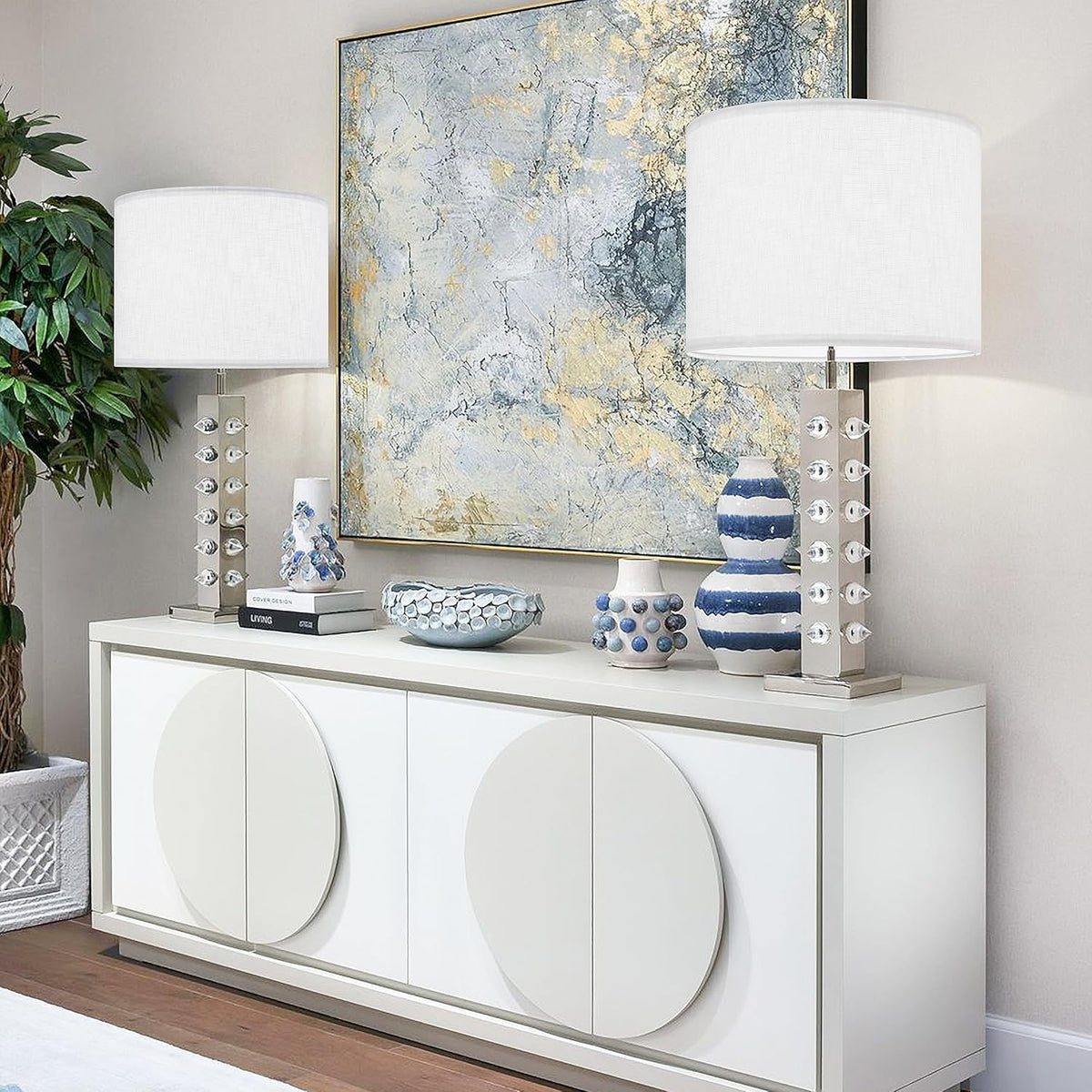 Two white drum lampshades on chrome studded table lamps flanking abstract art on a sideboard