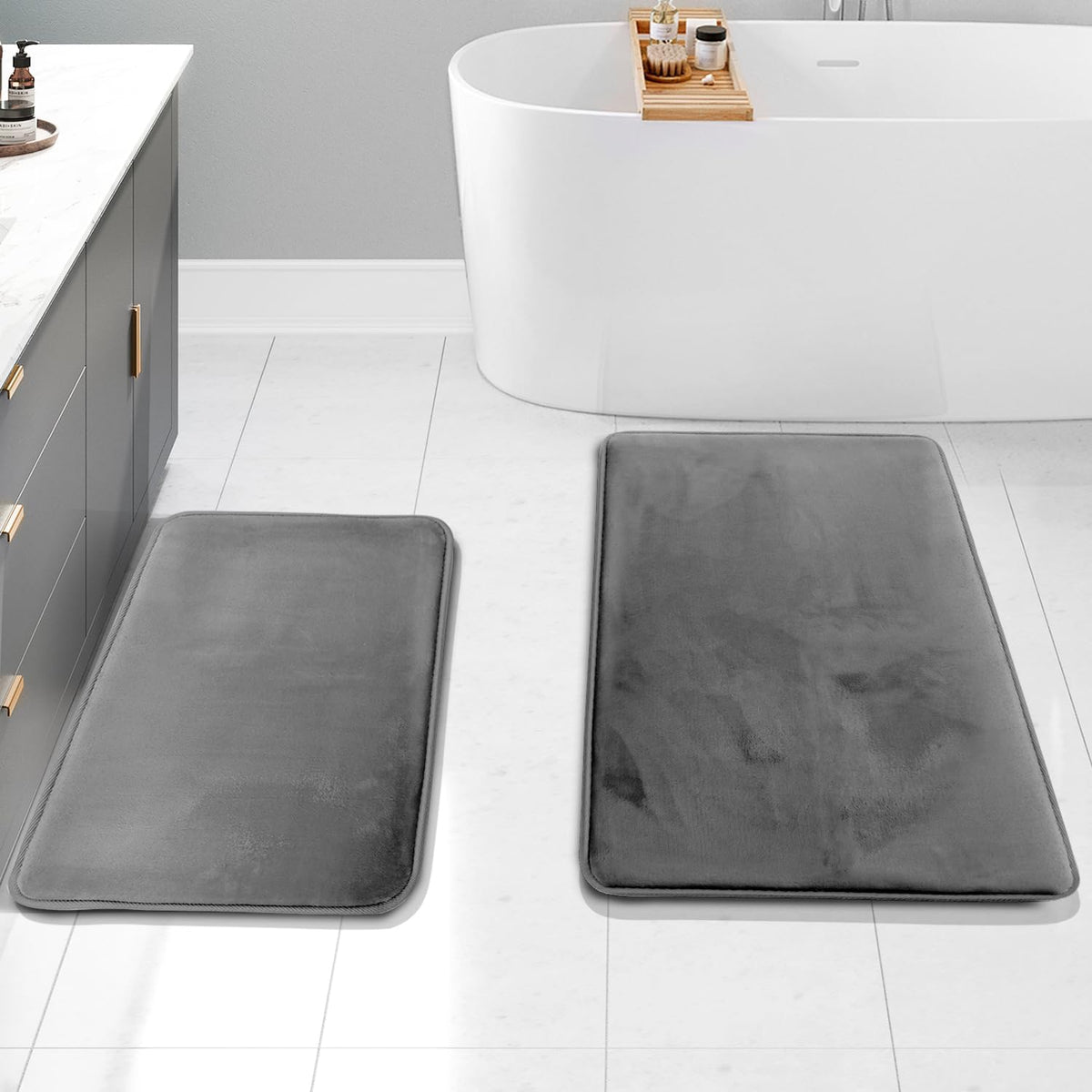 Two charcoal-gray rectangular memory-foam rugs placed on a light tile floor beside a freestanding tub and vanity.