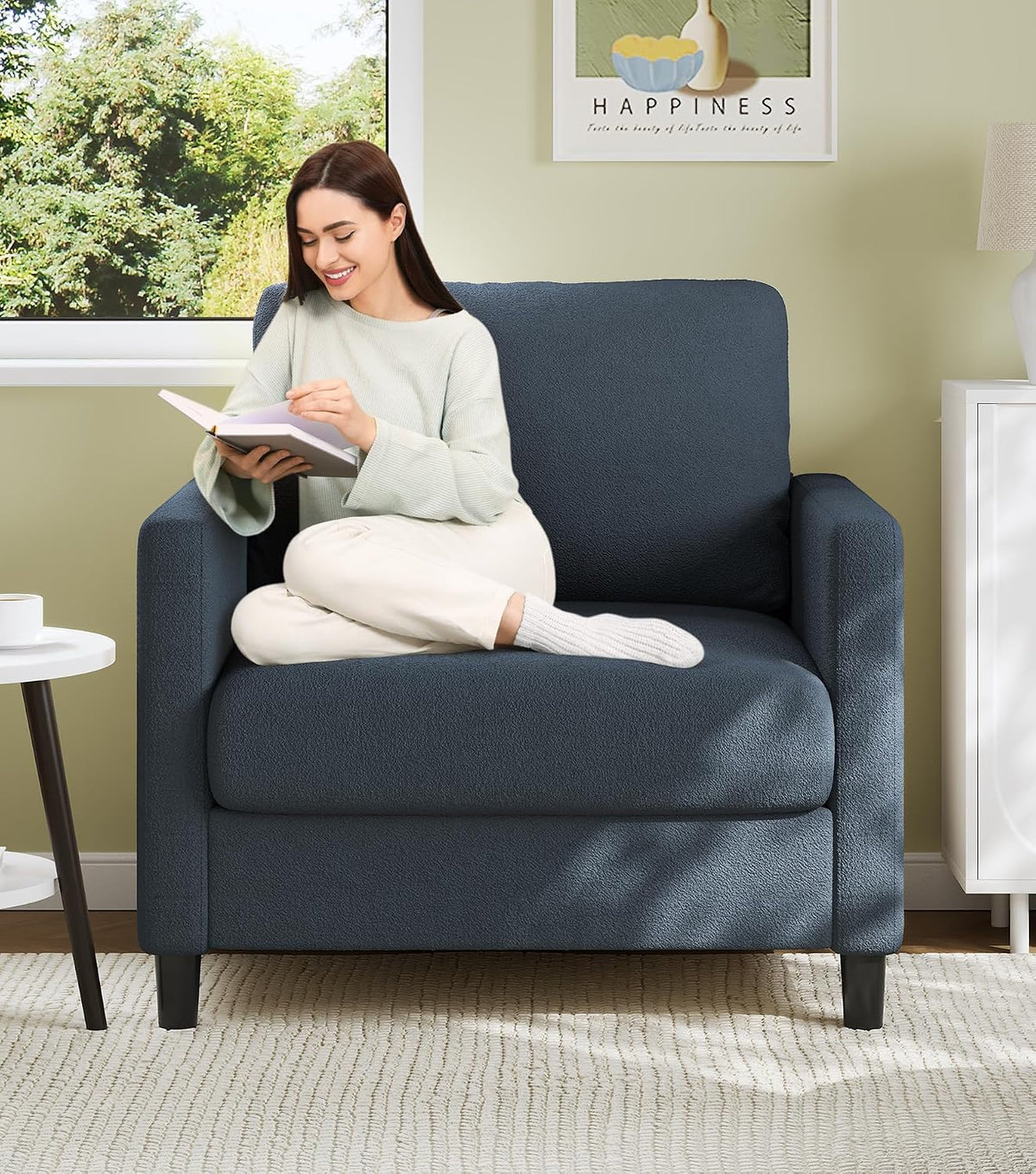 Woman reading on a small blue-gray teddy fabric loveseat near a window and side table.