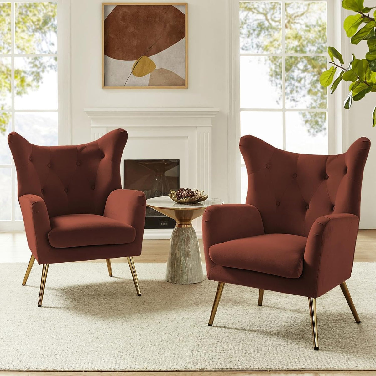 Two rust-colored tufted wingback chairs with gold-tone legs flanking a small round side table before a white fireplace.