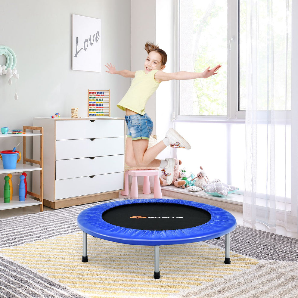 Blue round trampoline on a rug as a child jumps beside a dresser and bright windows.