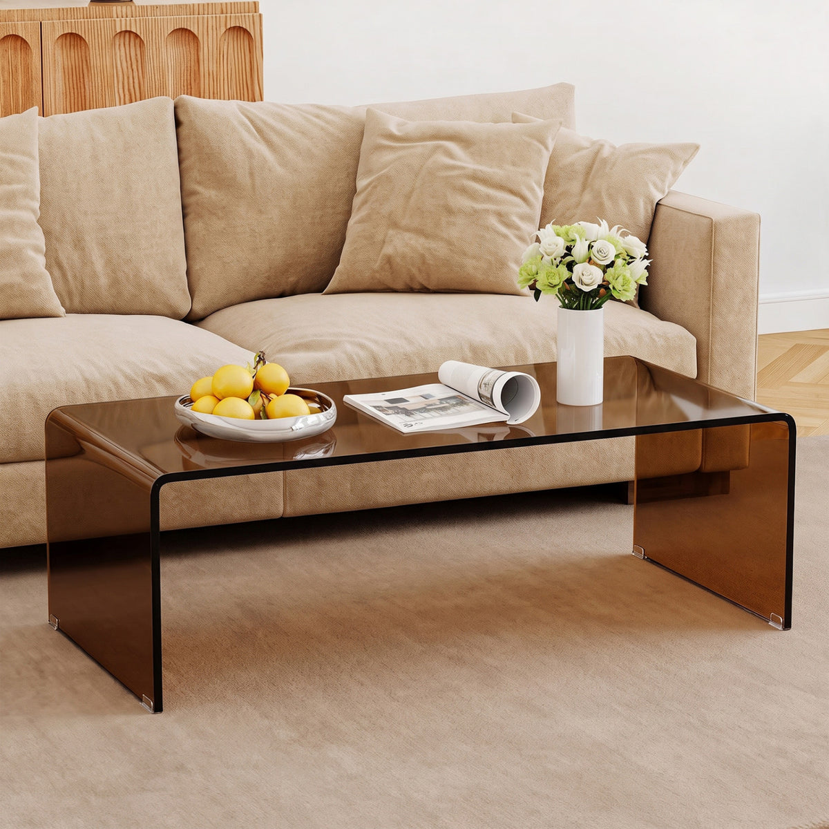 Dark glass coffee table in front of a beige sofa, topped with fruit, a magazine, and flowers.