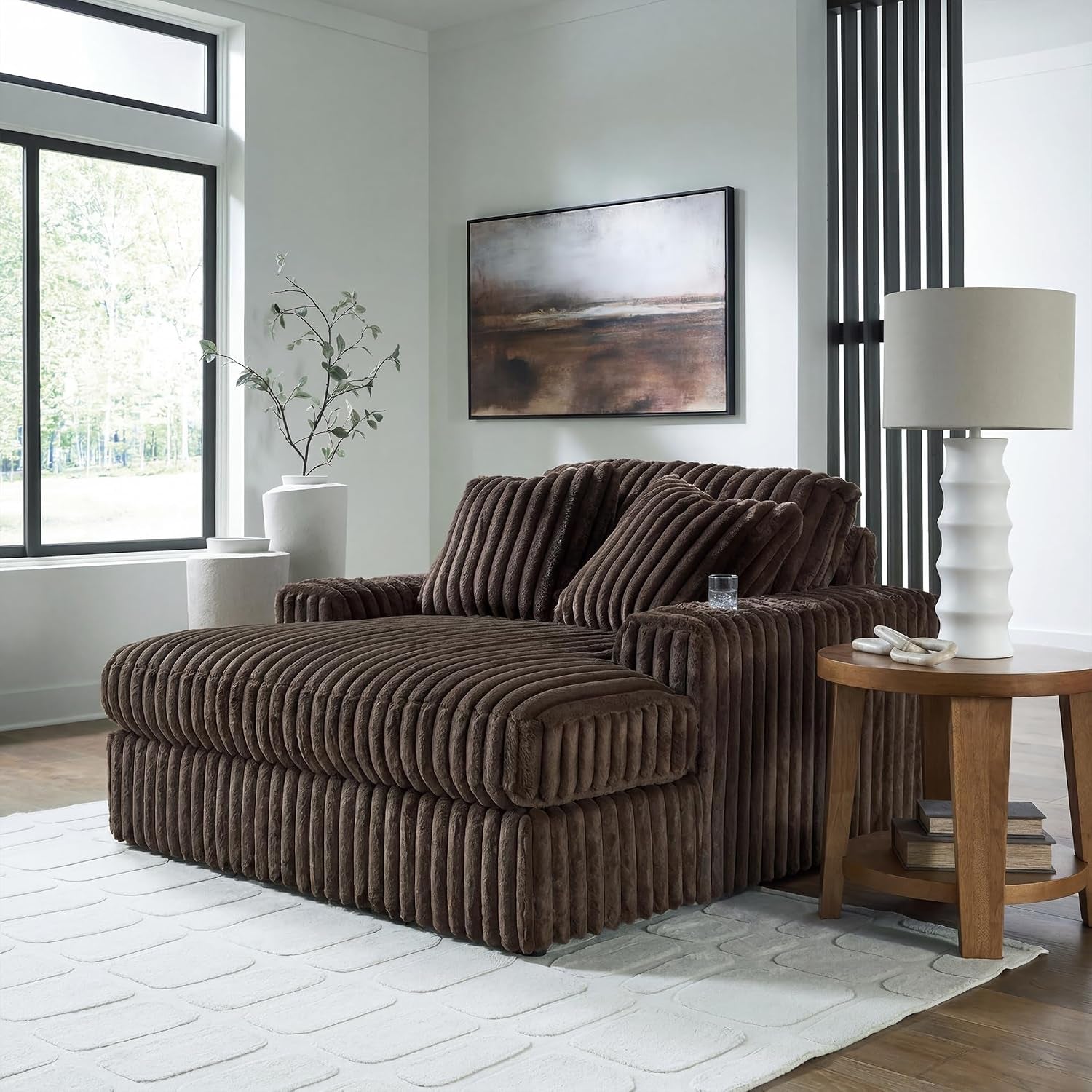 Brown chaise lounge staged in a bright living room beside a round wood side table and lamp with a glass on the arm