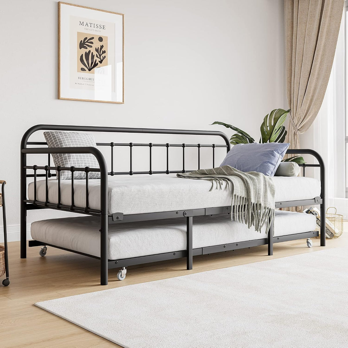 Black metal daybed with trundle pulled out showing two mattresses, caster wheels, pillows and a folded throw on top.
