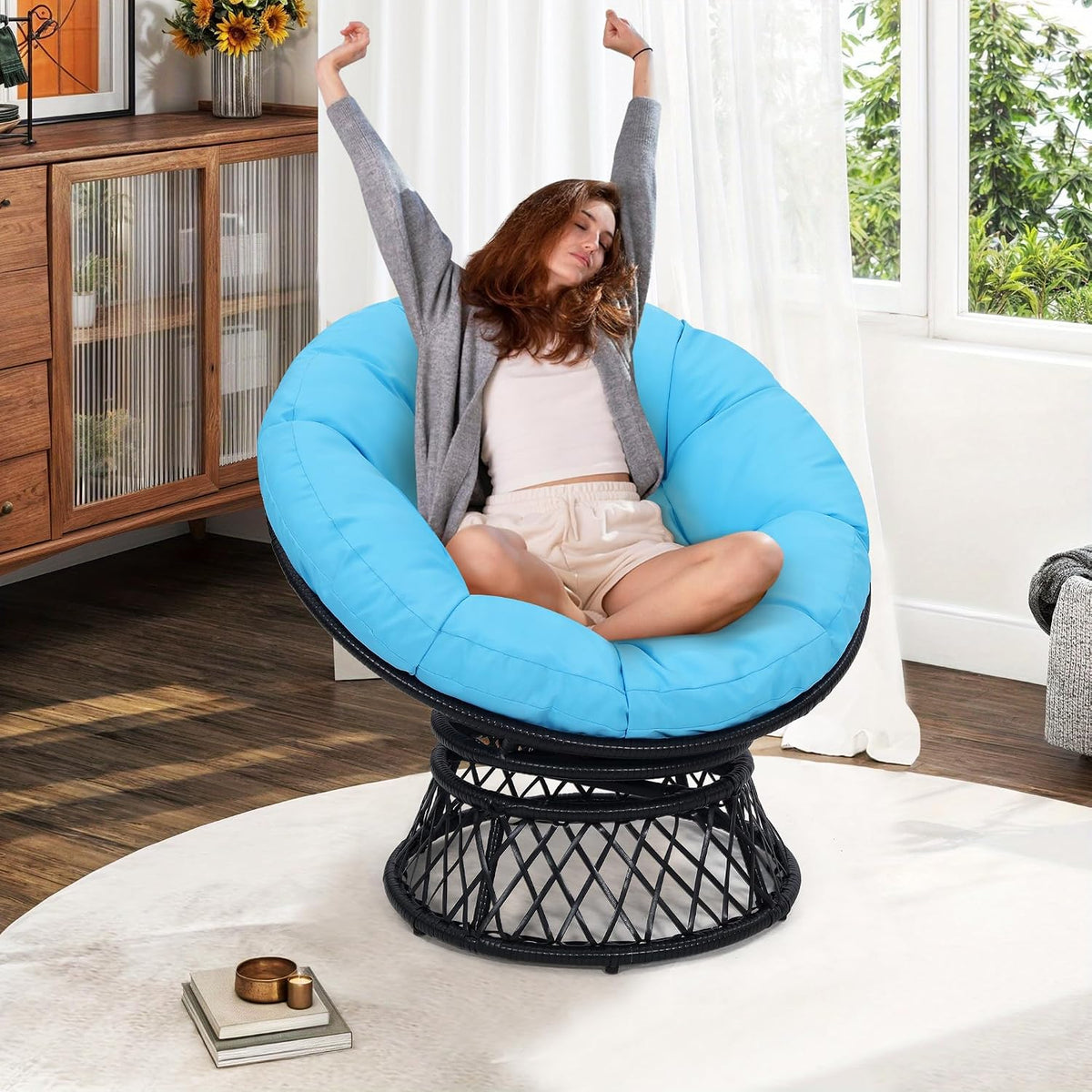 Same woman relaxing in a bright blue cushioned papasan chair with black woven swivel base near a window.