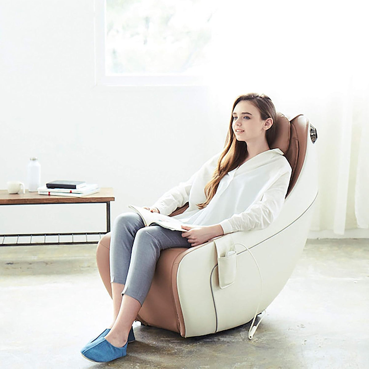Woman sitting in a beige massage chair in a bright room, holding a book with a side table behind her.