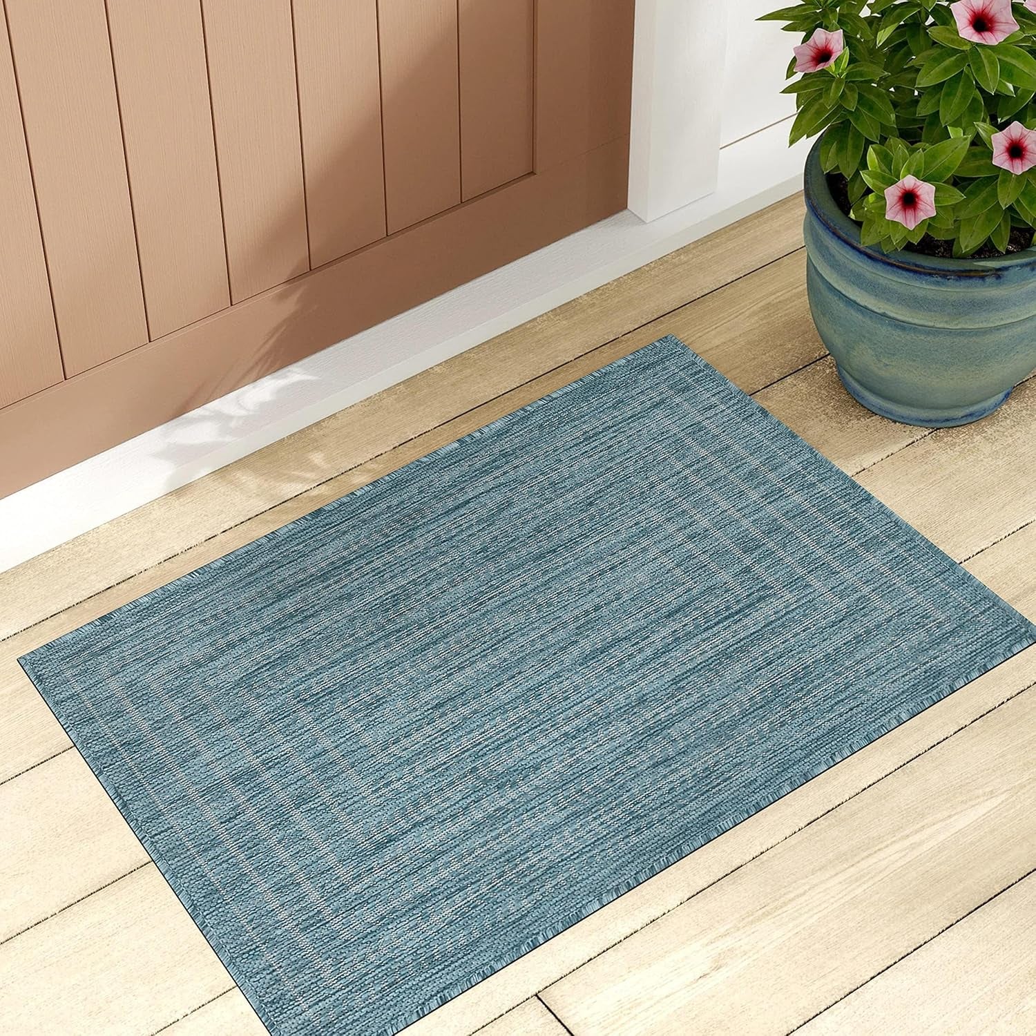 Teal textured rectangular rug on a wooden porch in front of a door, next to a blue planter with pink flowers.