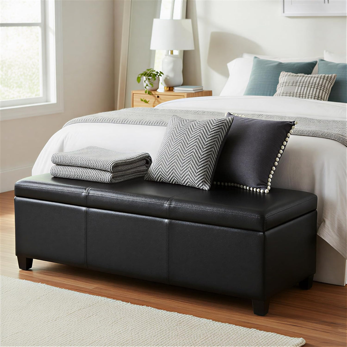 Black rectangular storage bench placed at the foot of a bed with pillows and folded throws on top.