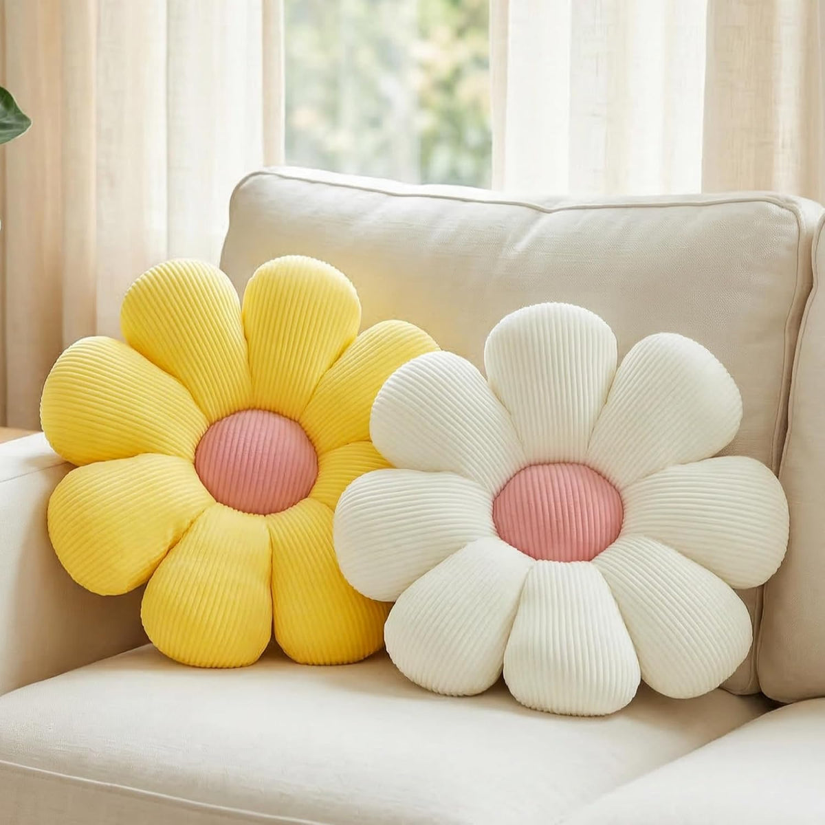 Close view of a yellow daisy pillow with pink center next to a cream daisy cushion on a beige sofa arm.