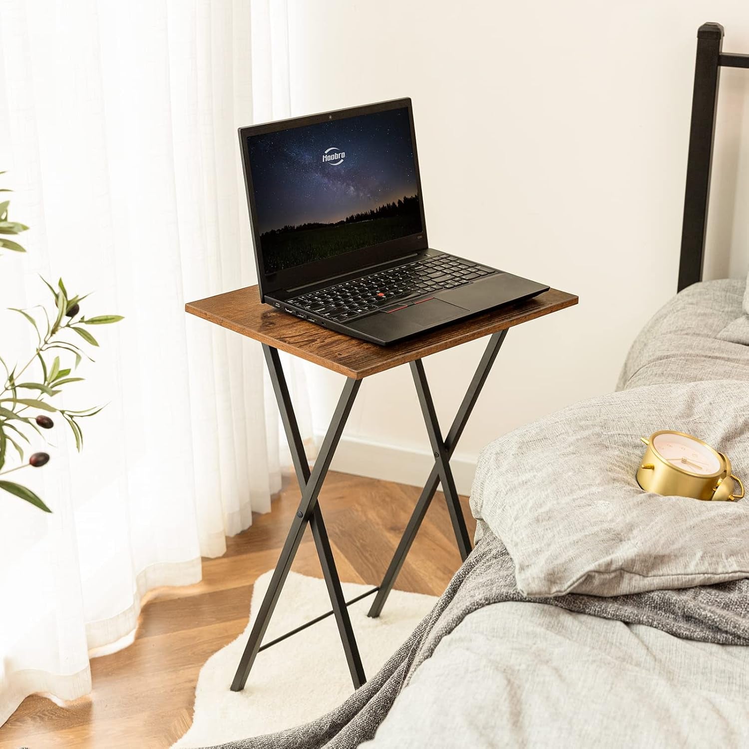 Rustic brown tabletop with a laptop on top and black metal X-legs visible beside a bed
