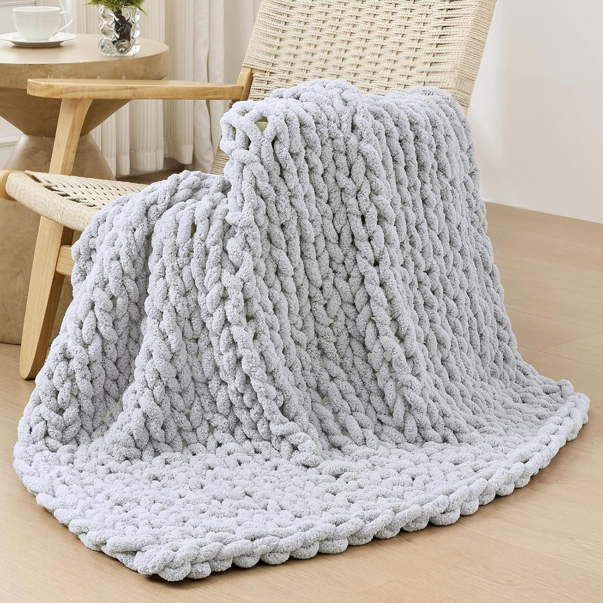 Light gray chunky knit throw draped over a woven chair, pooling onto a light wood floor in a bright room.