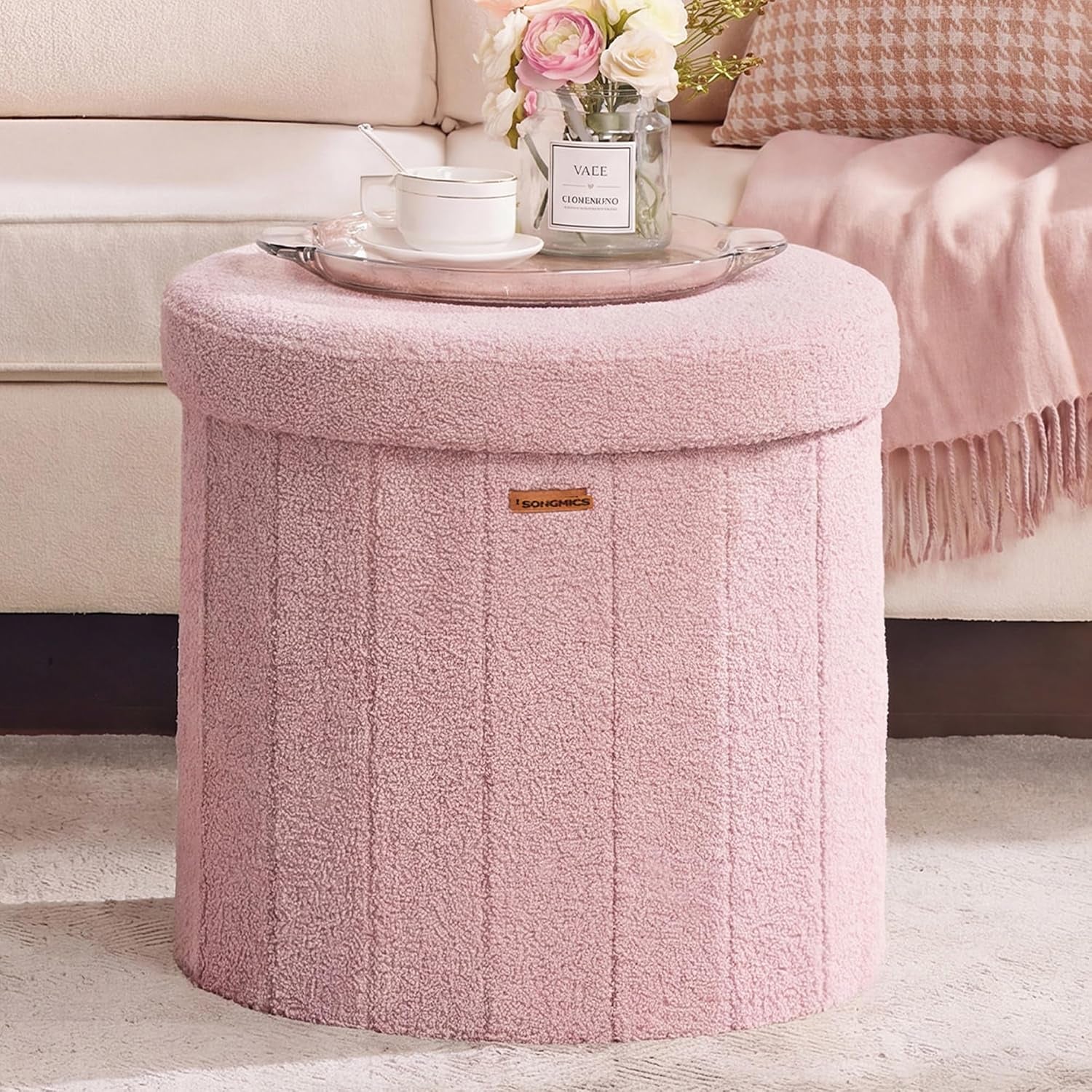Round pink plush storage ottoman with removable lid, clear tray and teacup on top beside a sofa.
