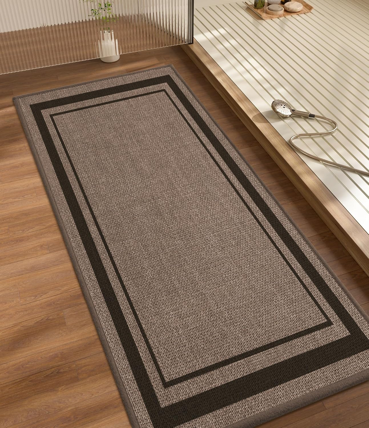 Long rectangular beige bath rug with double dark border on wood floor beside tub