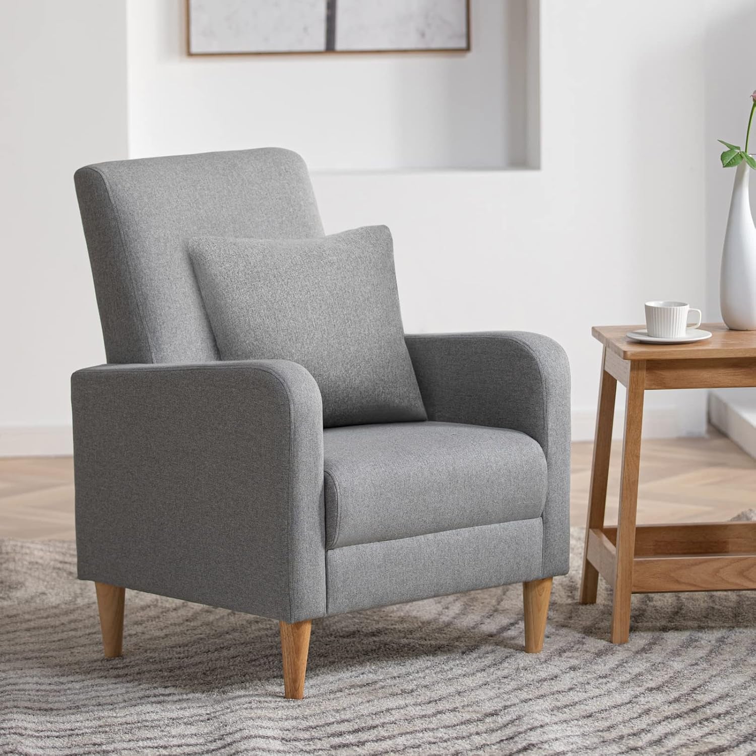 Light gray high-back accent chair with matching pillow beside a small wooden side table and rug.