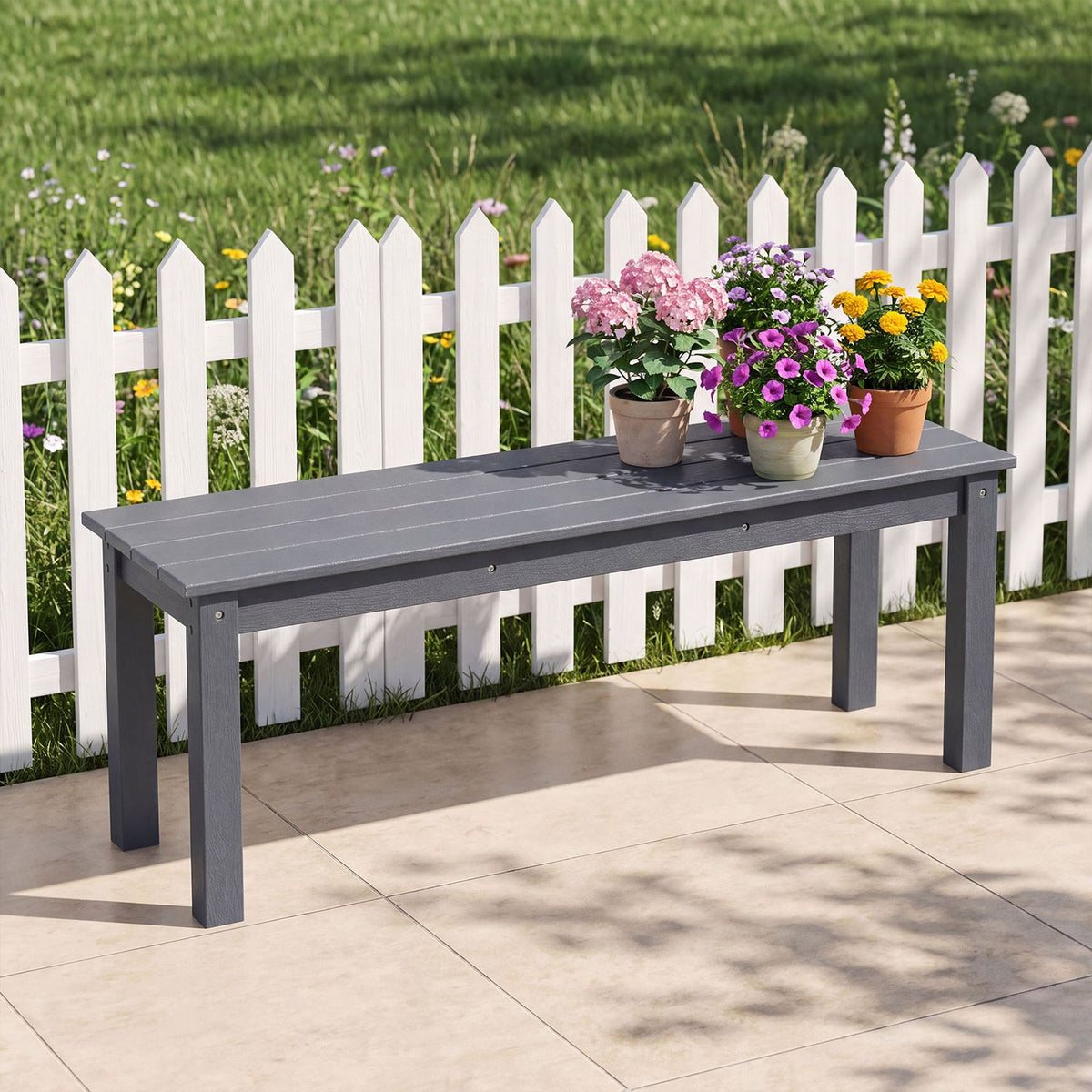 Dark gray slatted bench on a patio beside a white picket fence, topped with pots of flowers.