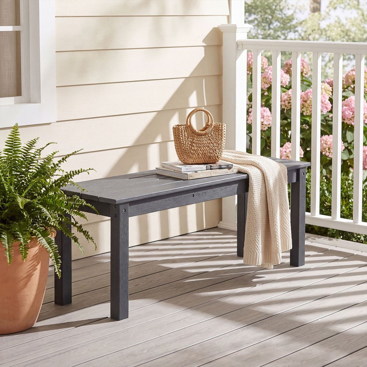 Dark slatted bench on a porch beside a potted fern, with a woven bag and throw draped over it.