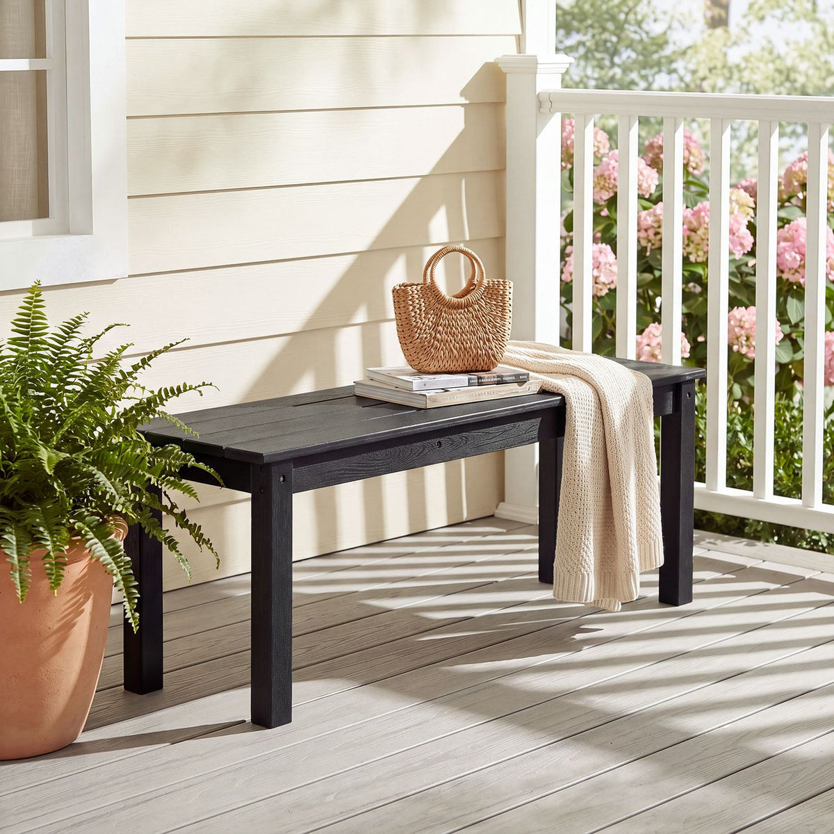 Black slatted bench on a porch beside a potted fern, with a woven bag and throw draped over it.