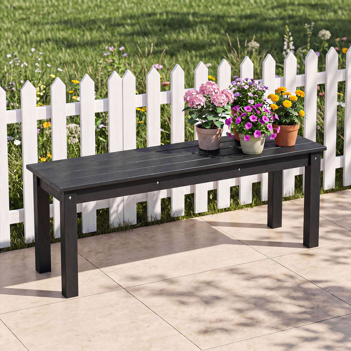 Black slatted bench with potted flowers, set on a patio in front of a white picket fence.