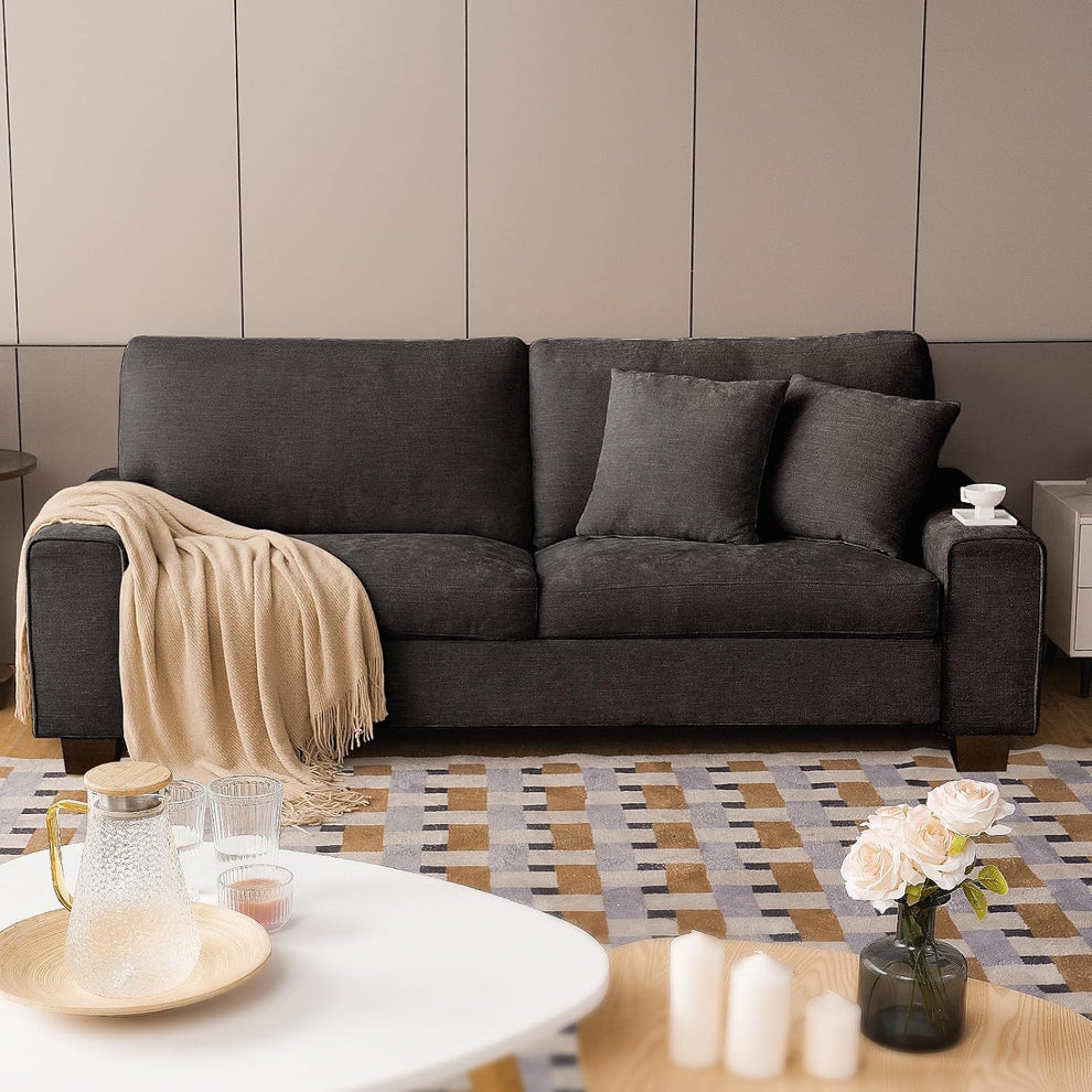 Dark gray loveseat shown with two throw pillows, a beige blanket over one arm, and a low white coffee table foreground