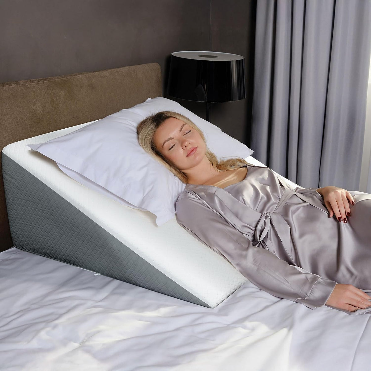 Top-side view of the wedge pillow under a reclining woman, showing the mattress edge and bedside lamp