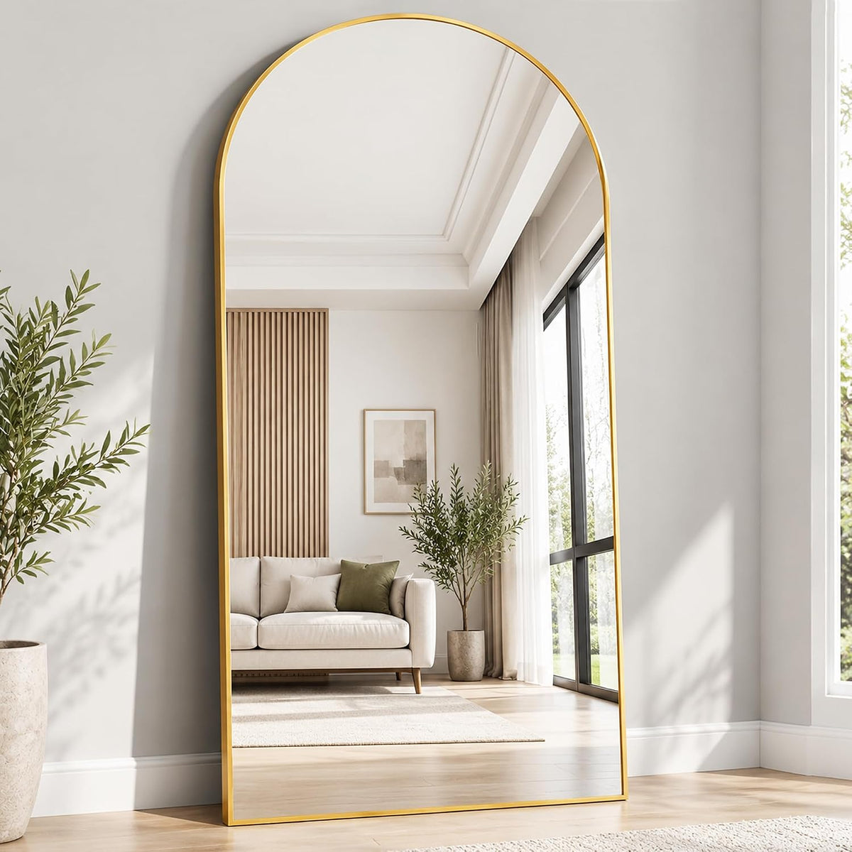 Arched full-length mirror with a gold frame standing on the floor reflecting a sofa and large windows.