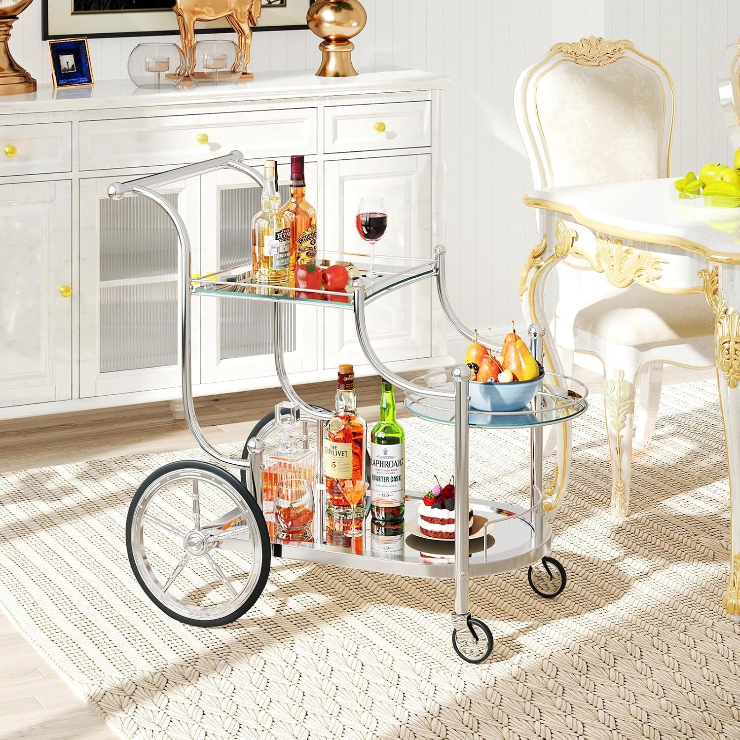 Silver three-tier glass bar cart staged with bottles, fruit bowl, and dessert in a dining room.
