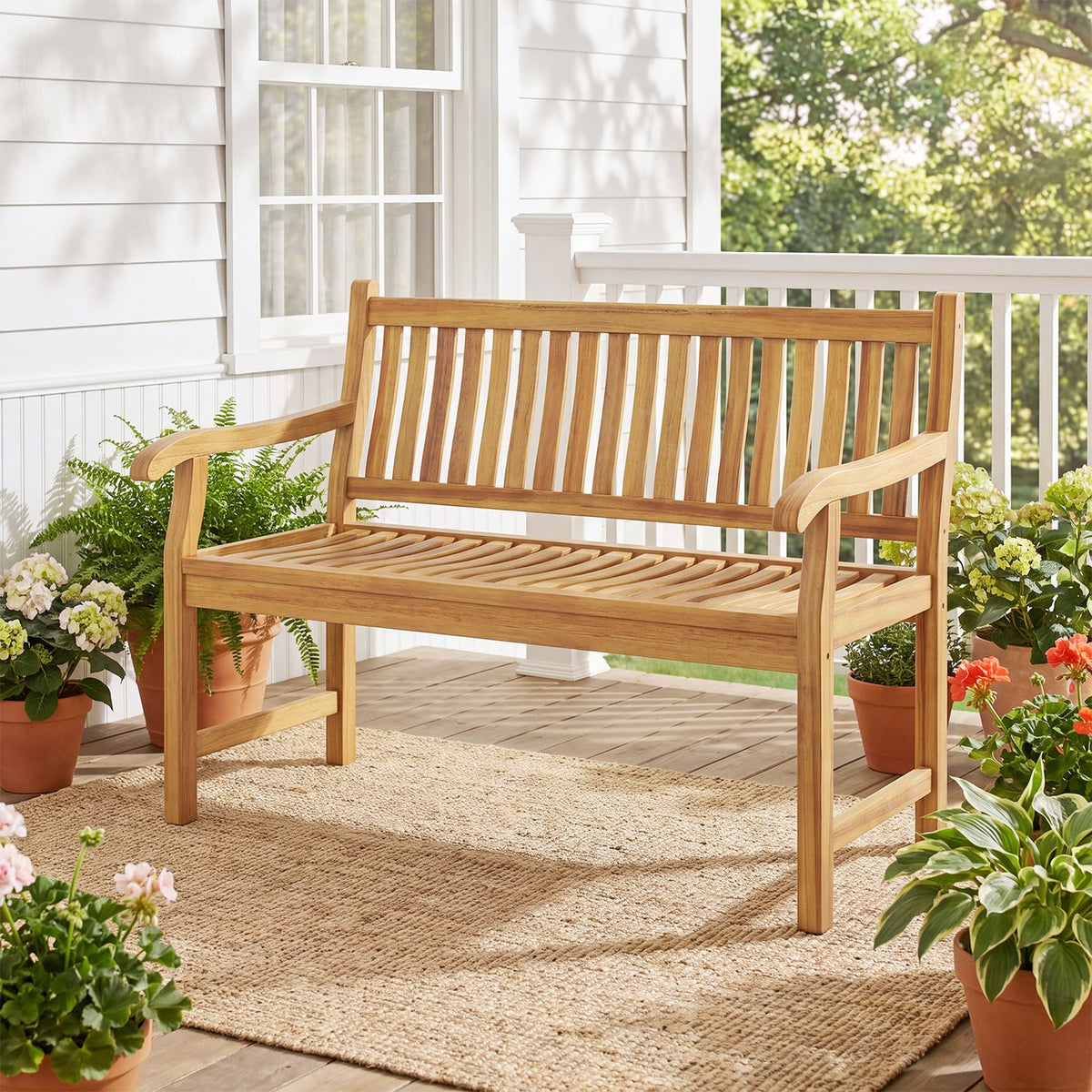Slatted teak-toned garden bench on a woven rug beside potted flowers on a sunny porch.