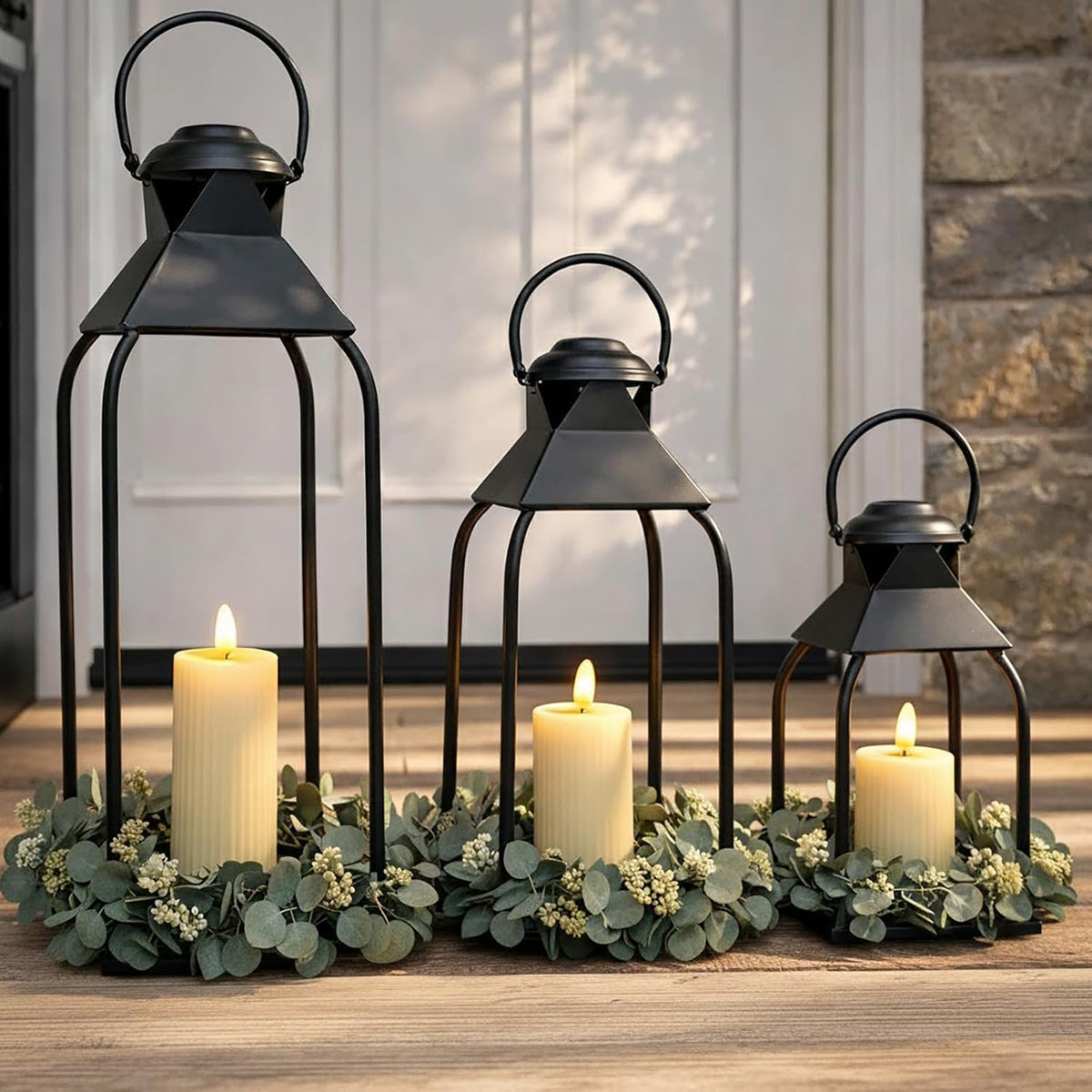Black metal open-frame lanterns in three sizes on a doorstep with pillar candles and greenery