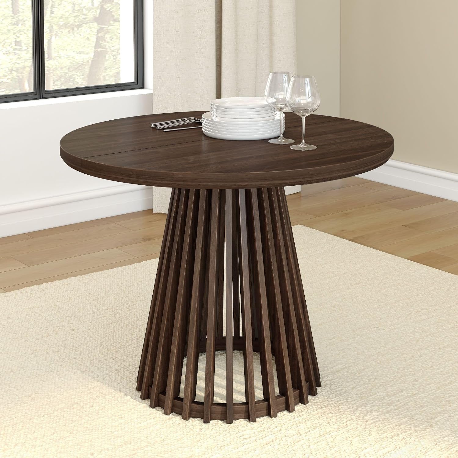 Round dark-wood dining table with slatted pedestal base, set with stacked plates and two wine glasses.