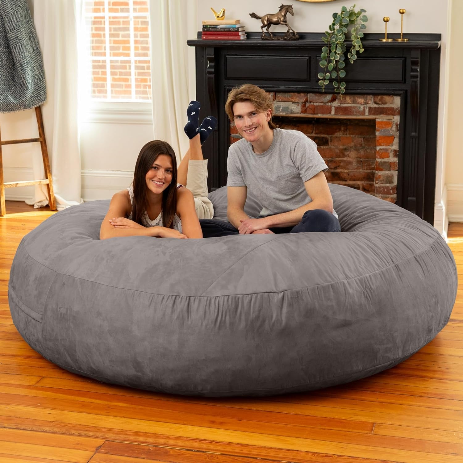 Large grey 6 ft bean bag bed in lounge mode with two people relaxing in a living room with a fireplace