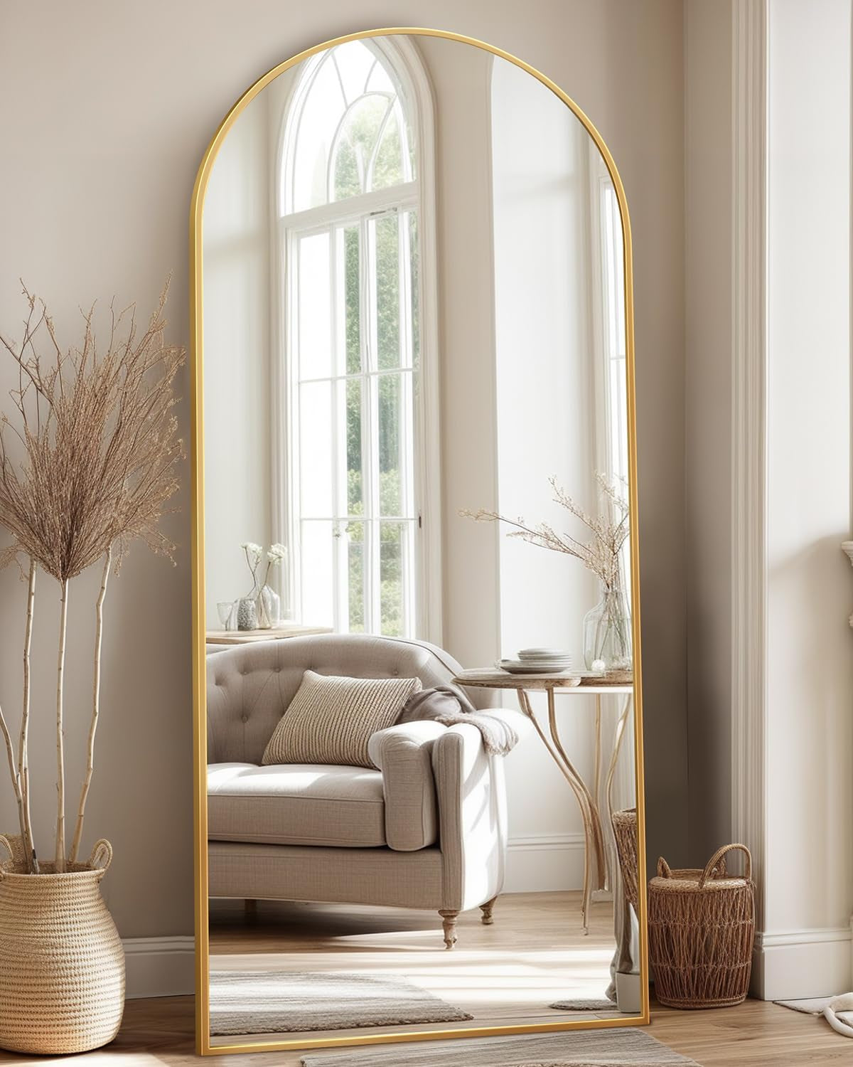 Large arched floor mirror with narrow gold frame propped against wall, reflecting an upholstered chair and window