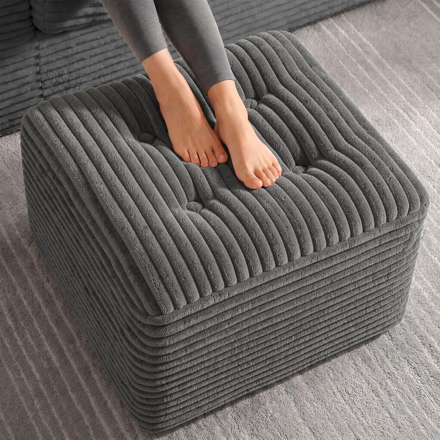 Close top-down view of a dark gray corduroy pouf with a person’s bare feet resting on it.