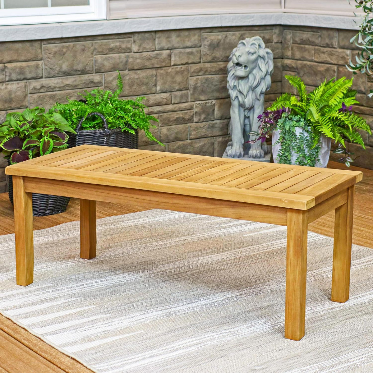 Teak coffee table on an outdoor rug in a patio setting with stone wall decor and potted plants in the background