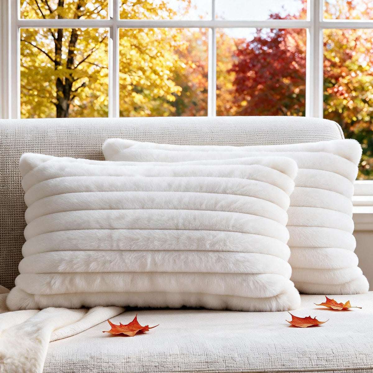 Two cream-white ribbed faux-fur square pillows arranged on a beige sofa in front of an autumn window.