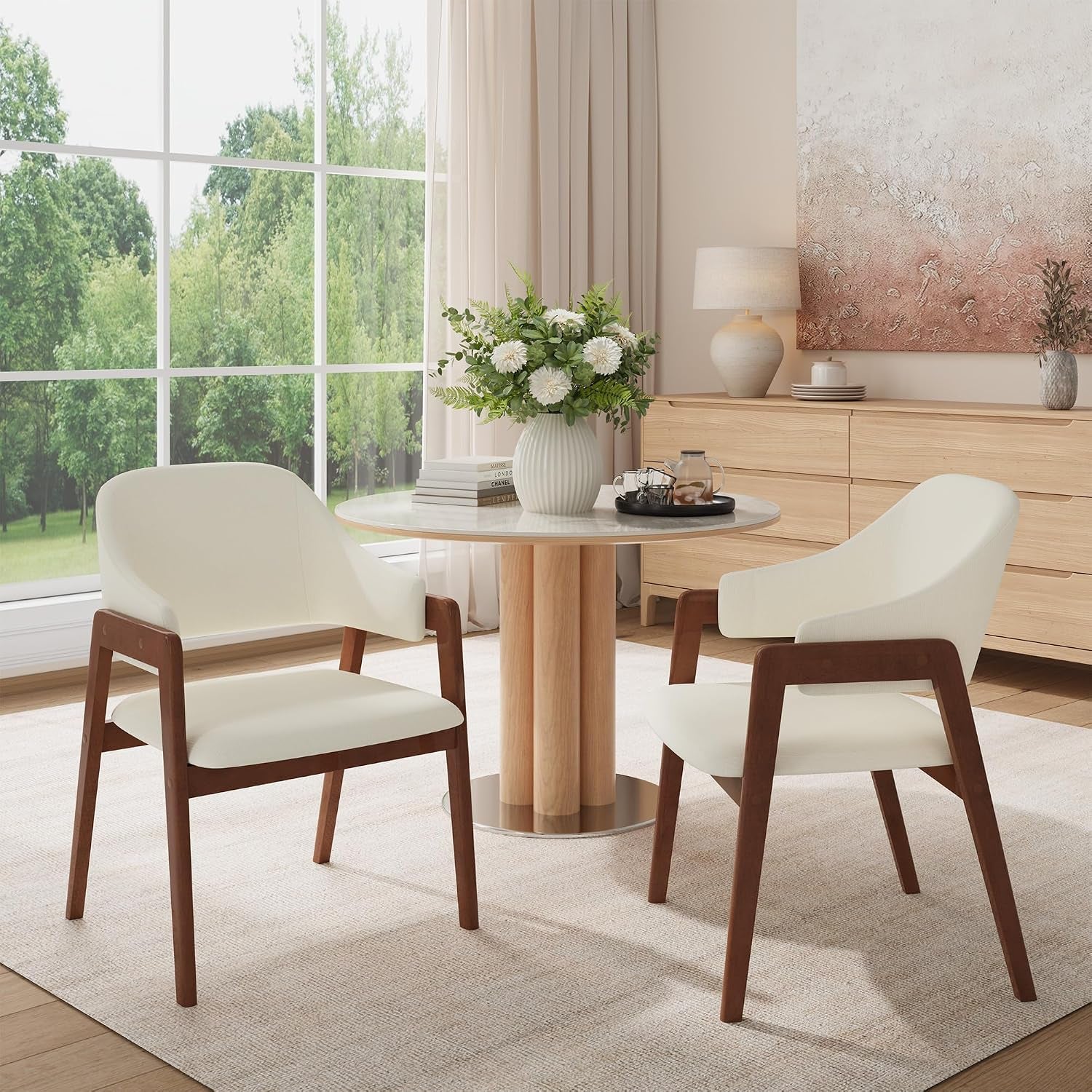 Two cream-upholstered chairs with walnut-tone frames flanking a small round table by a large window