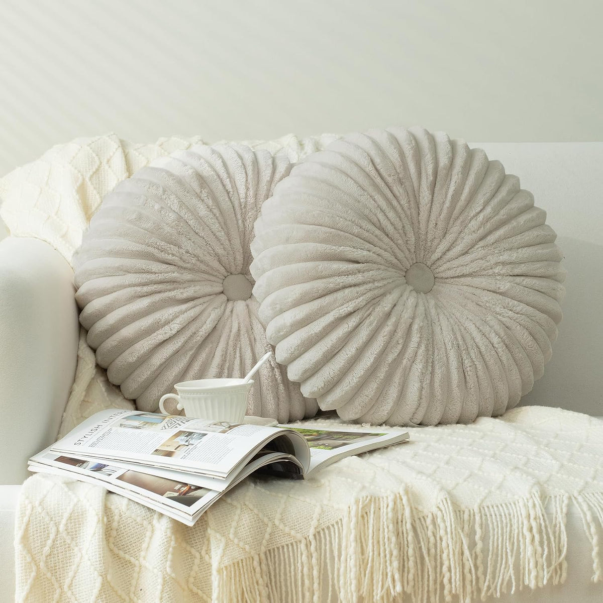 A pair of round ruched throw pillows in light beige on a sofa with an open magazine and teacup.