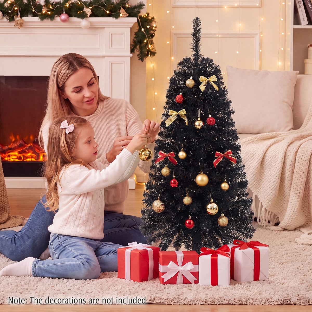 A woman and child decorate a dark green Christmas tree beside a fireplace and wrapped gifts.