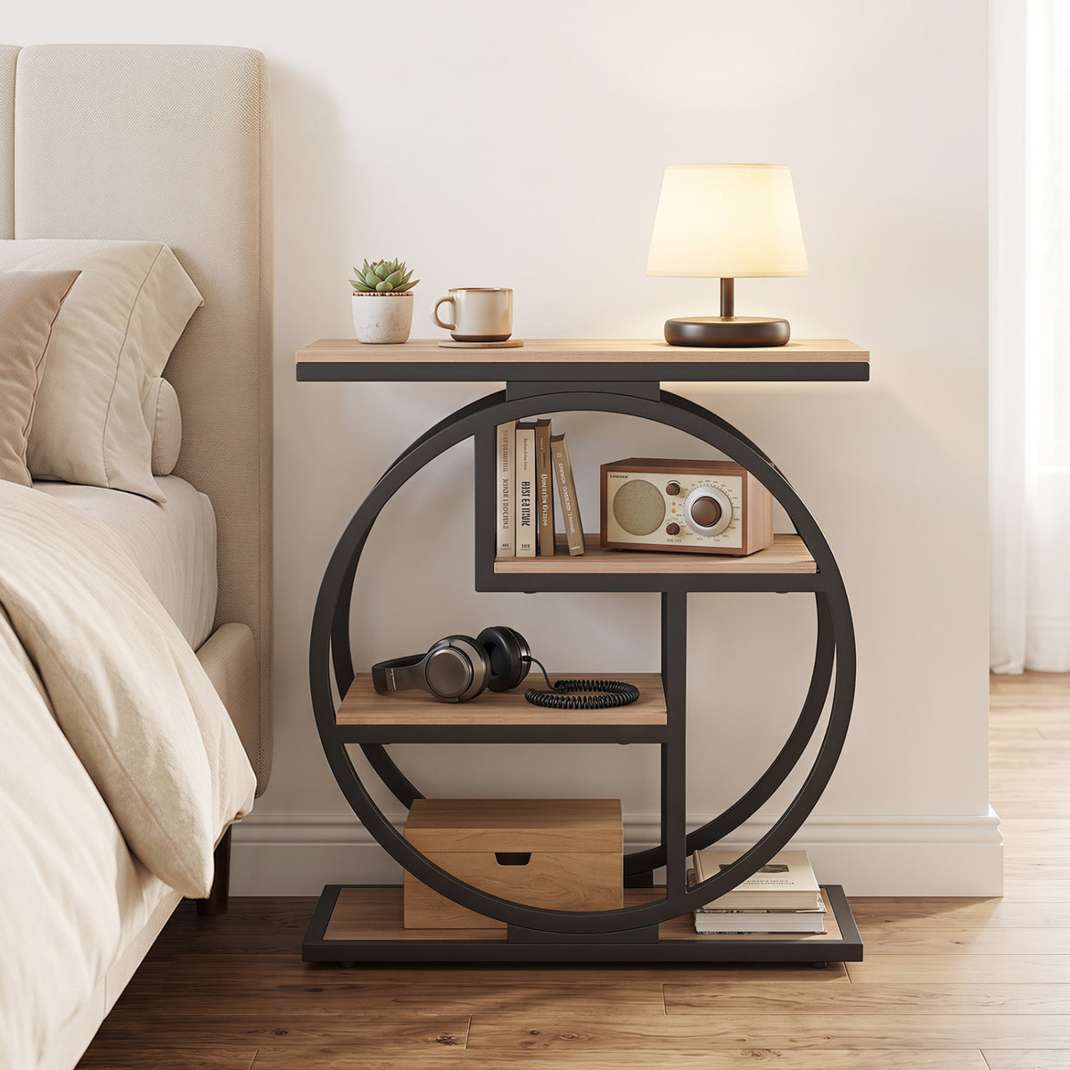 End table beside a bed, holding a small plant, mug, and lamp, with books and headphones on its open shelves.