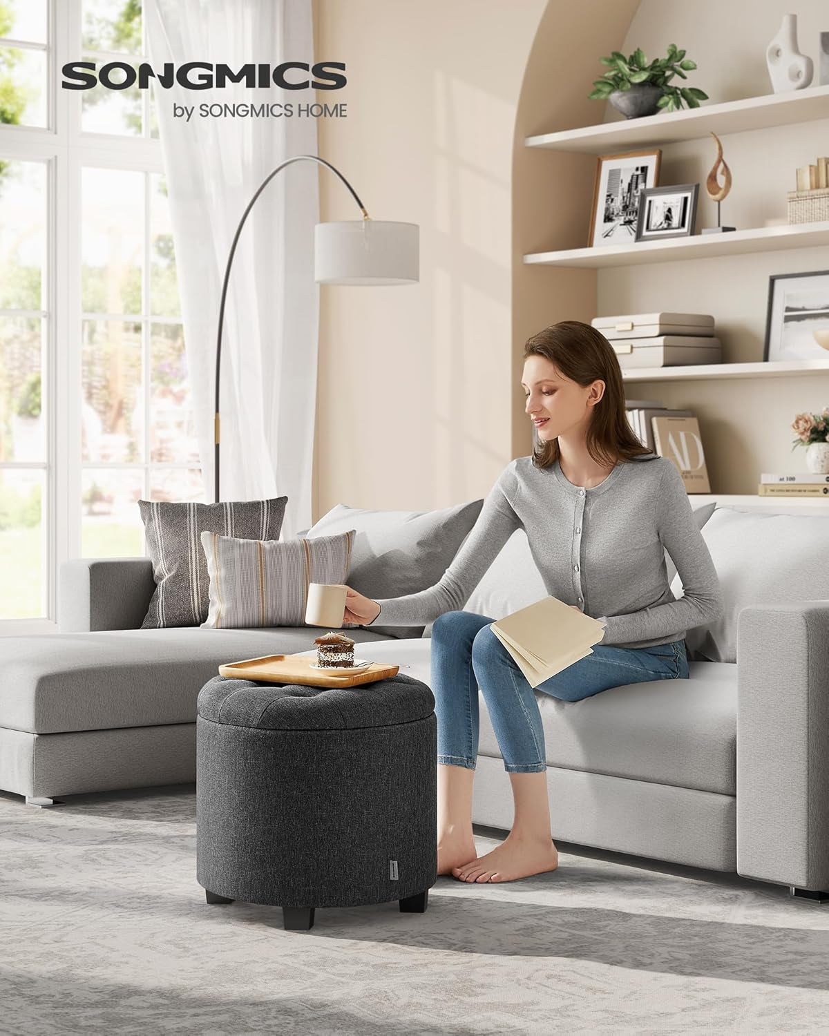 Dark gray round ottoman placed next to a sofa in a living room setting with a woman reading