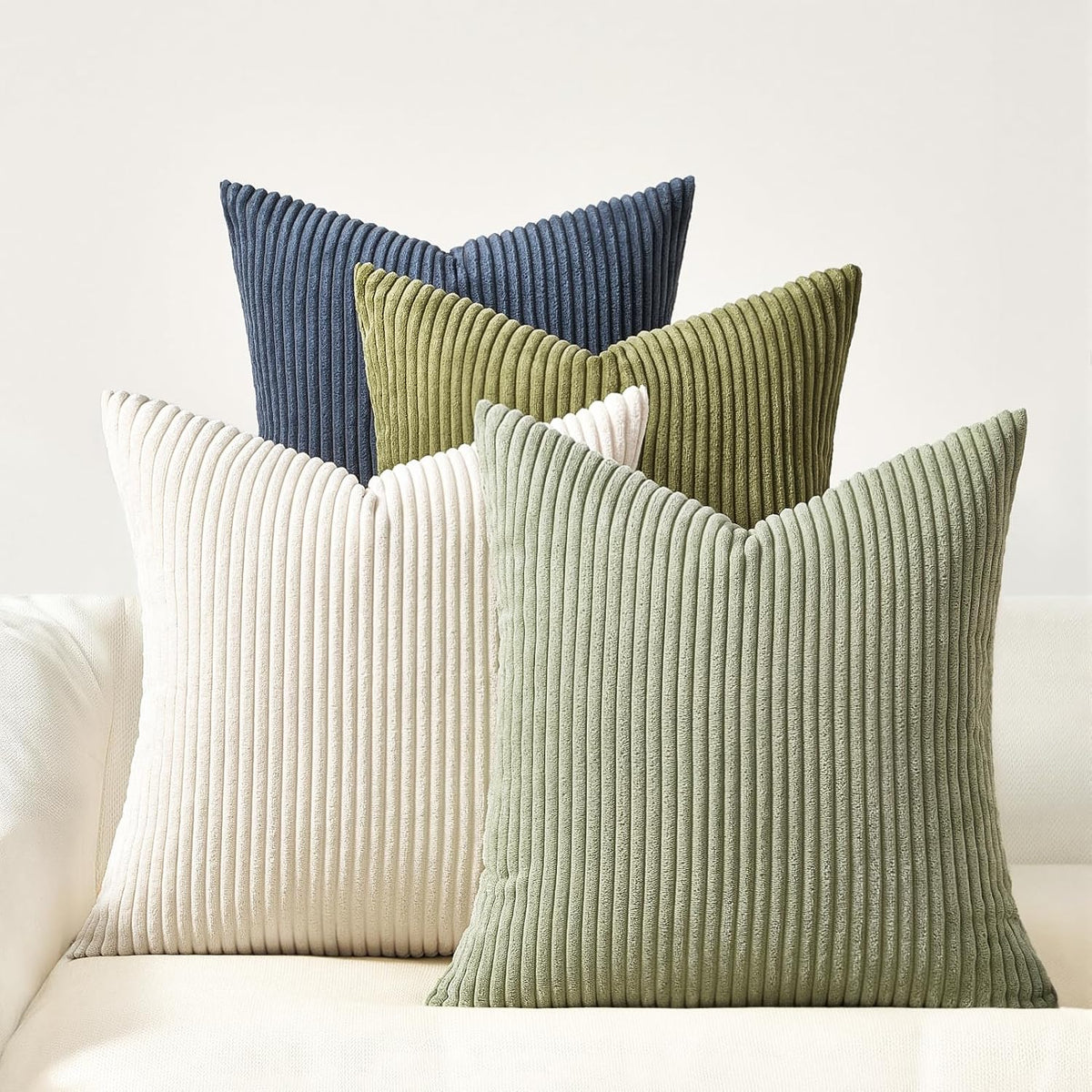 Five ribbed corduroy square pillows in blue, olive, sage and cream arranged on a light sofa.