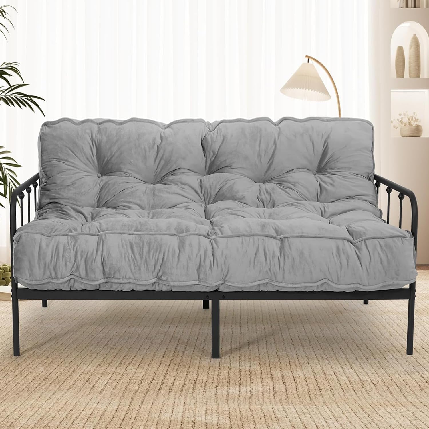 Full gray tufted futon mattress on black metal frame in a bright room with lamp and plants