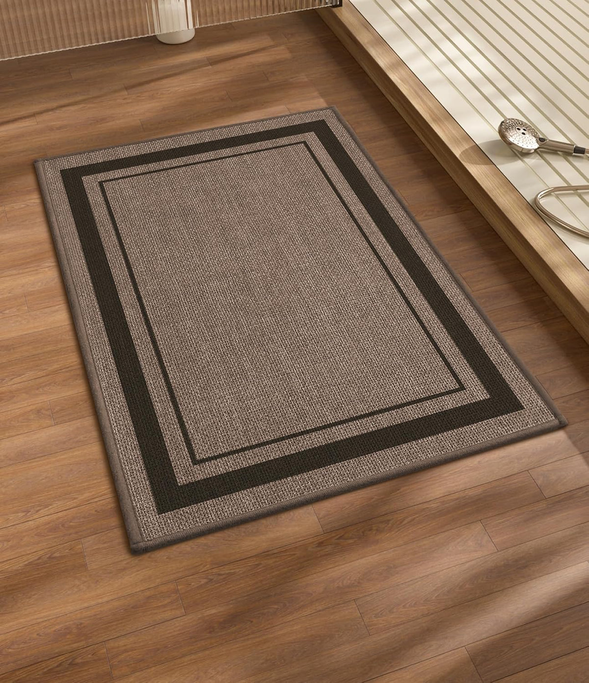 Brown rectangular bath mat with double-border on wood floor beside a raised shower platform and handheld showerhead.