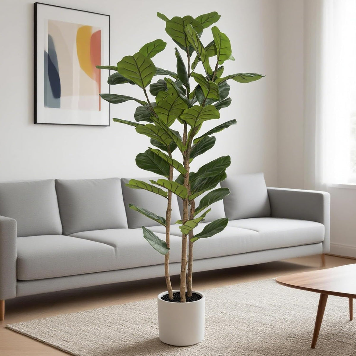 Faux fiddle leaf fig in a white pot standing on a beige rug in front of a gray sofa and framed wall art.