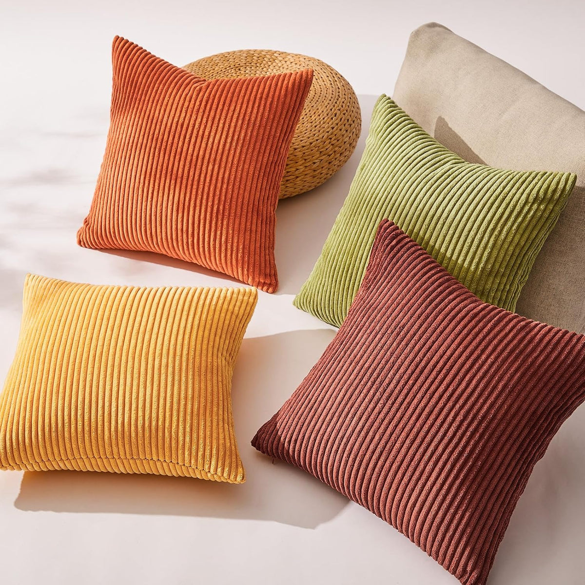 Four ribbed corduroy pillows arranged on a light surface with a woven pouf and a sofa corner, showing fabric texture