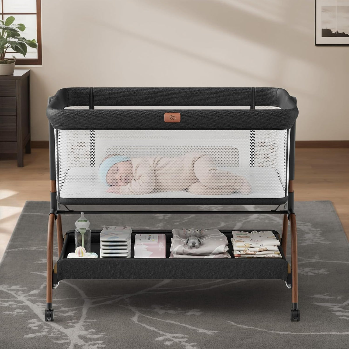 Dark gray/black bedside bassinet with mesh sides, sleeping baby inside and organized shelf below.