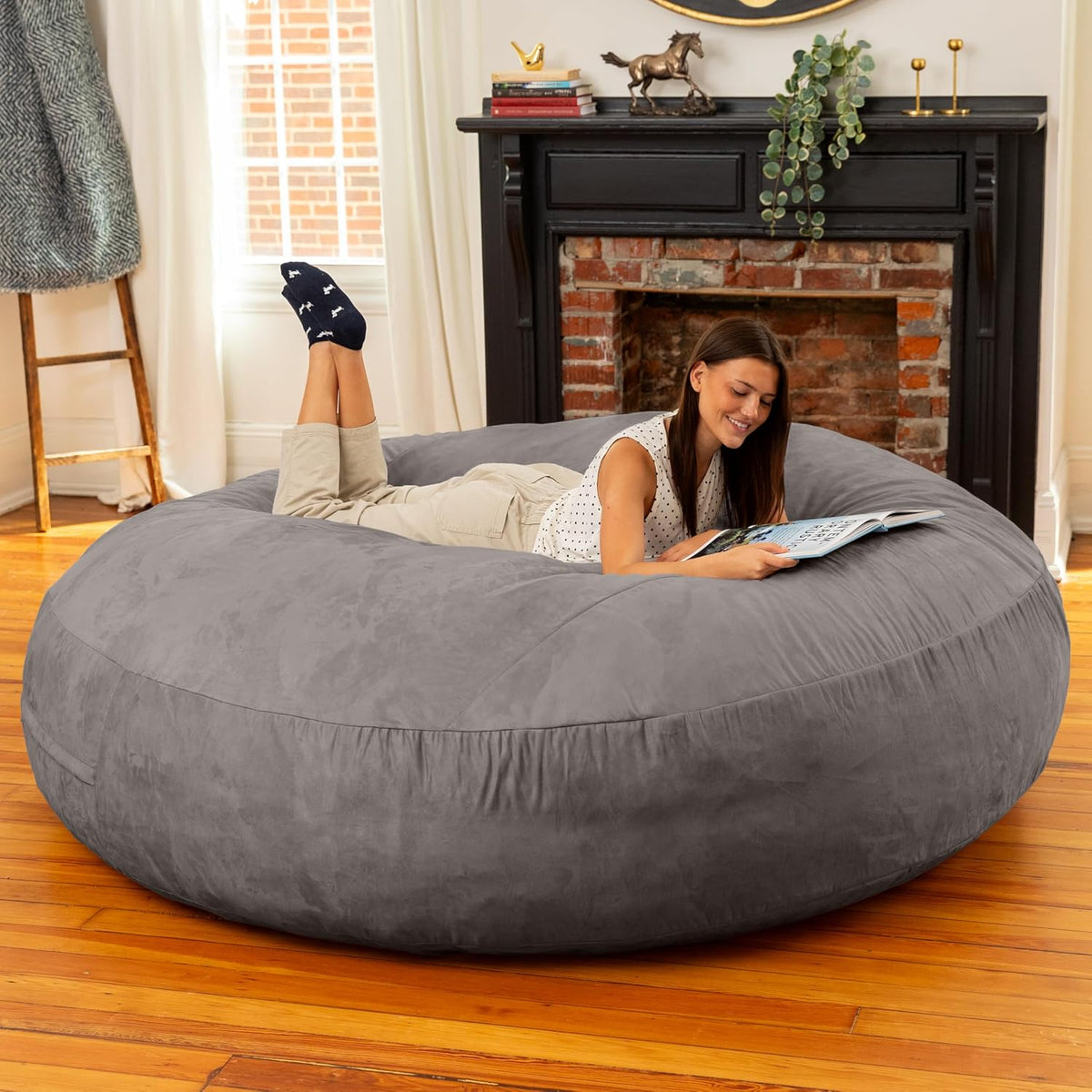 Woman lying on her stomach reading on a large gray round bean bag in a living room in front of a fireplace.