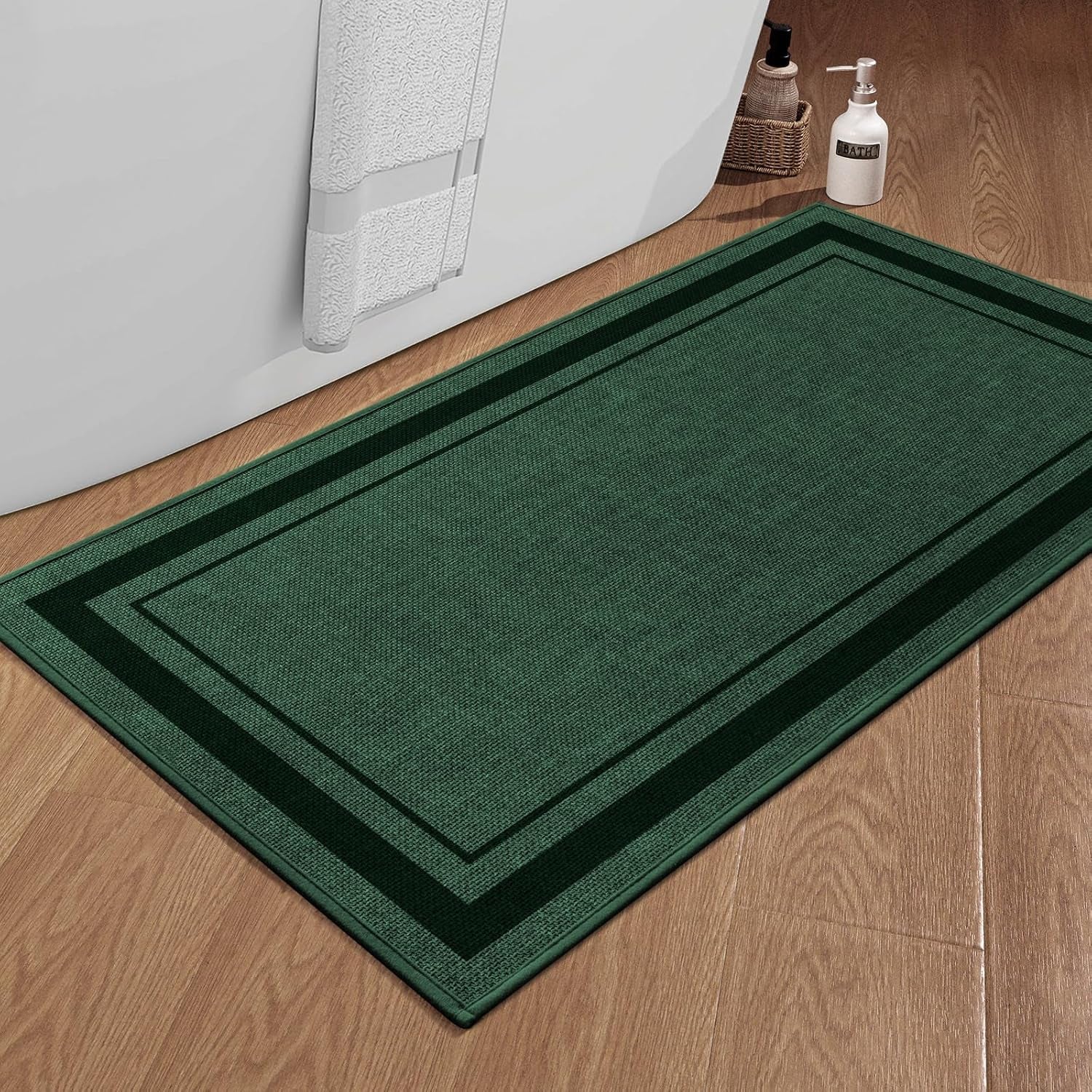 Forest green rectangular bath mat with darker double border positioned next to a bathtub on wood floor