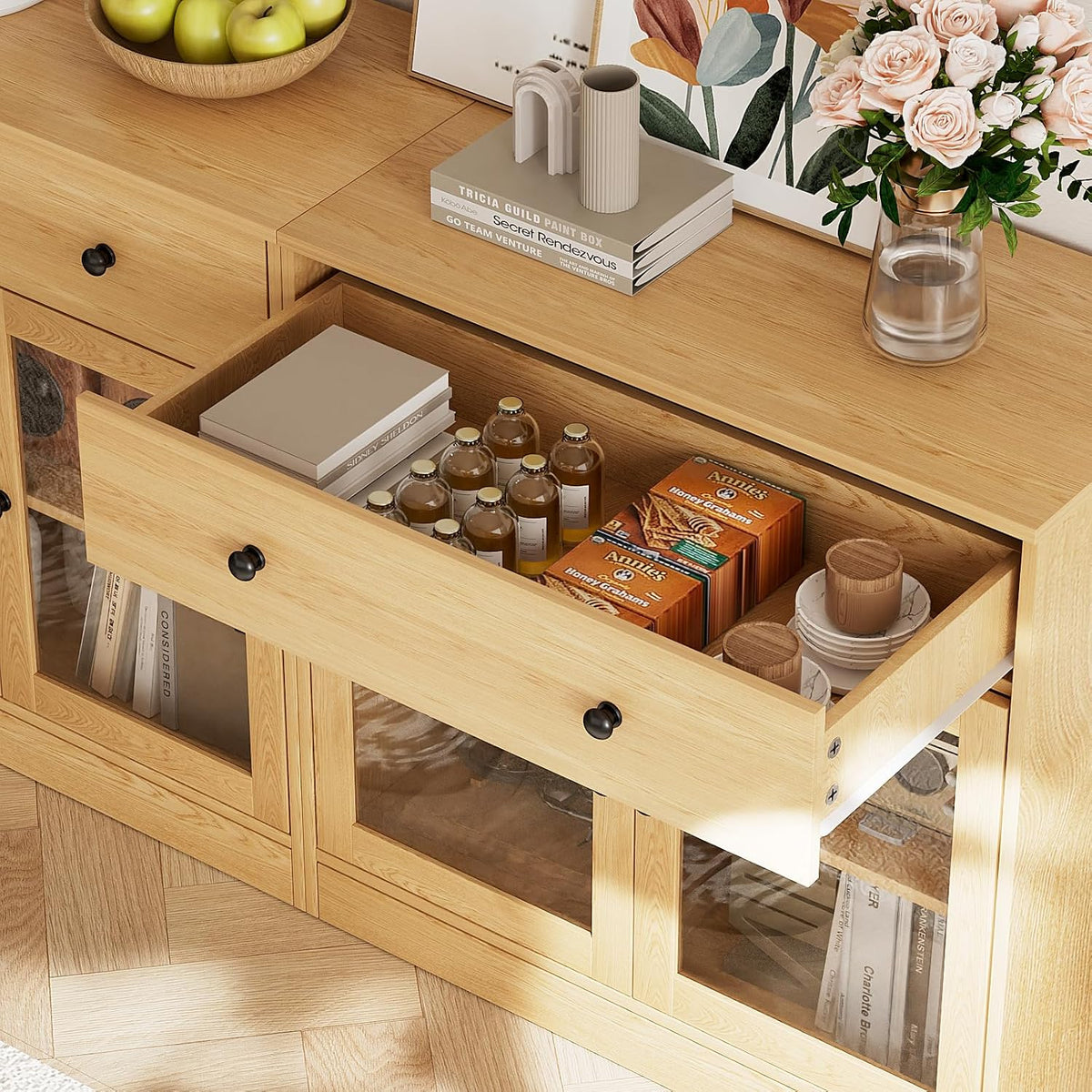 Top drawer pulled open showing bottled drinks, boxed snacks, stacked plates and books inside, with decor on the unit top.